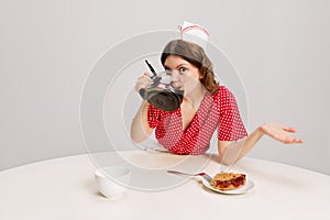 Stylish young female waitress in retro american fashion style of 70s, 80s sitting at table and drinking coffee over grey