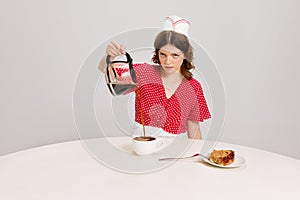 Stylish young female waitress in retro american fashion style of 70s, 80s sitting at table and drinking coffee over grey