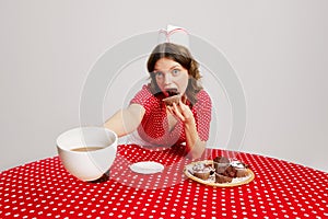 Stylish young girl in retro american fashion style of 70s, 80s uniform sitting at table and tasting cupcakes with coffee