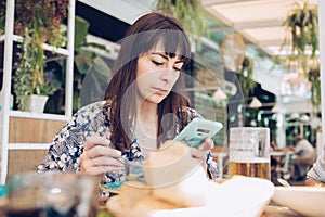 Stylish young girl with a serious face looking at her smart phone while eating at a restaurant