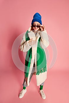 Stylish young girl in sunglasses, blue hat, green scarf and fur coat posing over pink background. Comfortable outfit