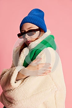 Portrait of stylish young girl in sunglasses, blue hat, green scarf and fur coat posing over pink background. Feeling