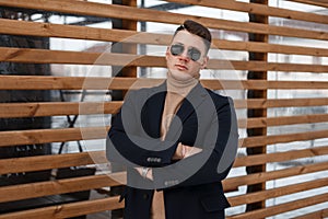 Stylish young hipster man in a fashionable black coat in a warm knitted sweater in trendy sunglasses is posing near a wall