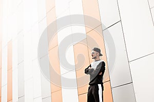 Stylish young hipster man in stylish cap in sunglasses in trendy black sports suit is standing near a modern building in the city