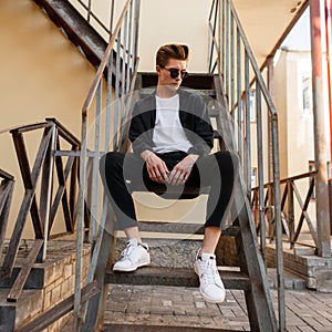 Stylish young hipster man in trendy sunglasses in a black shirt in a t-shirt in elegant striped pants in sneakers sits