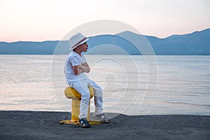 Kid in white clothes sitting by the ocean, sea with mountains on background. Sitting and waiting on seascape and