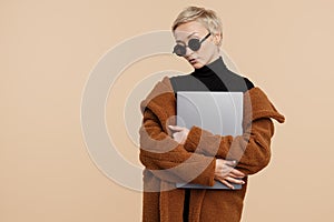 Stylish young hipster woman with blonde short hair wearing a coat and sunglasses holding computer over beige background.