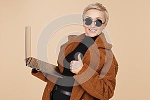 Stylish young hipster woman with blonde short hair wearing a coat and sunglasses holding computer over beige background.