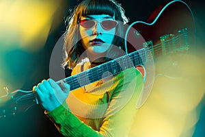 Stylish young hipster woman with curly hair holding red guitar on shoulder in neon lights. 90s style concept.