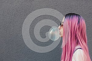 Stylish young hipster woman with long pink hair blowing a bubble with bubble gum.