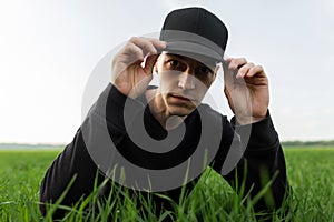 Stylish young man in a black shirt in a black trendy cap adjusts his cap and rests in green grass in the field. Handsome guy model