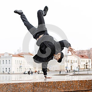 Stylish young man dancer in black jeans in a stylish jacket in a cap in sunglasses does a handstand in the city on the street