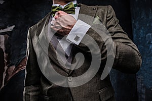 A stylish young man dressed in an elegant suit correct his bow tie against a dark textured wall