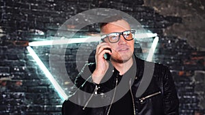 Stylish young man with glasses talking on mobile phone on background shabby brick wall with glowing lamps. Portrait of