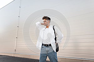 Stylish young man with hairstyle in white fashionable clothes with trendy hairstyle with bag posing near a modern building