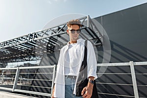 Stylish young man hipster in dark sunglasses in white fashionable clothes with hairstyle with black bag stands near a gray