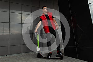 Stylish young man hipster in a trendy red vest in sunglasses in black jeans stands next to a vintage scooter near a black building