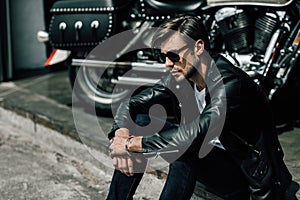 Stylish young man in leather jacket and sunglasses sitting on concrete curb near motorbike