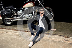 Stylish young man in leather jacket and sunglasses sitting on concrete curb near motorbike