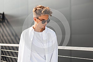 Stylish young man model in vintage white shirt in trendy sunglasses with a hairstyle stands near a gray building on a sunny day.