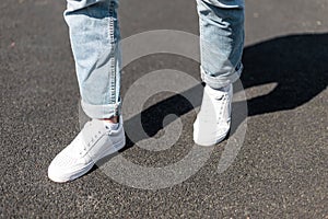 Stylish young man stands on an asphalt road in leather white sneakers in stylish blue jeans. Fashionable men`s shoes.