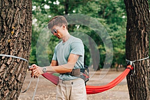 Stylish young man ties a hammock in the woods to a tree, ties the slings with his hands. The process of tying a hammock in the