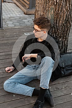 Stylish young man tourist in fashionable jeans wear in glasses with leather black backpack is resting near tree on street. Cool