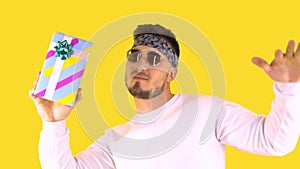 Stylish Young Man In Trendy Glasses, Bandana Holds A Gift And Dances. Back yellow background. Festive mood