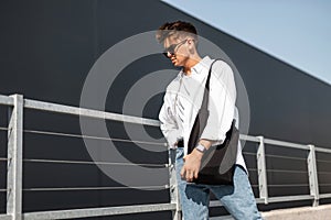 Stylish young man in vintage white shirt in fashionable sunglasses trendy blue jeans with a cloth black bag posing on a sunny day