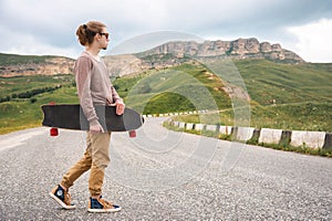 A stylish young man walks along a winding mountain road with a skate or longboard in his hands the evening after sunset