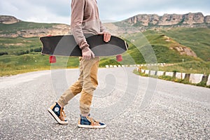 A stylish young man walks along a winding mountain road with a skate or longboard in his hands the evening after sunset