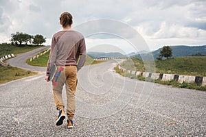A stylish young man walks along a winding mountain road with a skate or longboard in his hands the evening after sunset