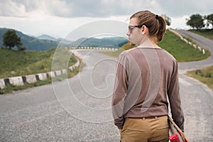 A stylish young man walks along a winding mountain road with a skate or longboard in his hands the evening after sunset