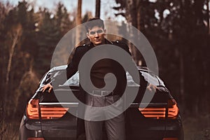 A stylish young man wearing a black coat leaning on a luxury car in the autumn forest