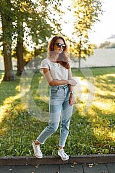 Stylish young redhead hipster woman in vintage jeans in a white T-shirt in trendy sunglasses in stylish sneakers posing in a park