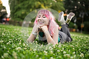 Stylish young smiling woman with multicolored pink hair lying on green grass in city summer park