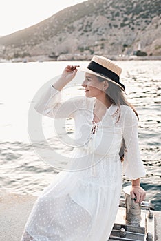 Stylish young woman 24-26 year old wear straw hat and white elegant dress posing over sea at quayside outdoors.