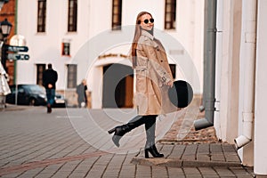 Stylish young woman in a beige coat and a black hat in his hands and glasses on a city street. Women`s street fashion. Autumn