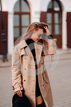 Stylish young woman in a beige coat and a black hat in his hands and glasses on a city street. Women`s street fashion. Autumn