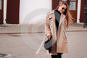 Stylish young woman in a beige coat and a black hat in his hands and glasses on a city street. Women`s street fashion. Autumn