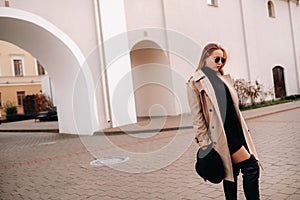 Stylish young woman in a beige coat and a black hat in his hands and glasses on a city street. Women`s street fashion. Autumn