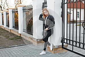 Stylish young woman in a black spring jacket in trendy jeans in a white polo shirt in sneakers posing near a vintage metal fence.