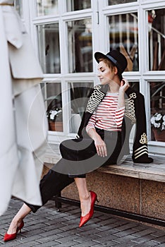Stylish young woman blonde in a hussar jacket, black hat, jeans, bananas and red shoes in the city near the window of the