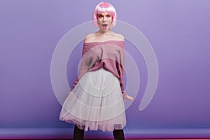 Stylish young woman in bright pink periwig standing on purple background. Inspired good-looking girl in peruke and long
