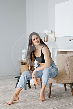 Stylish young woman in casual home clothes, jeans and a black top, sitting on a chair in the living room