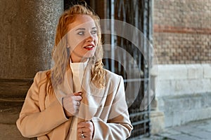 Stylish young woman in coat stands near the column smiles and looks away. Girl with red curly hair outside