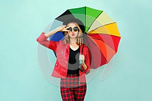 Stylish young woman with colorful umbrella blowing red lips sending sweet air kiss, wearing red jacket, black hat on blue wall