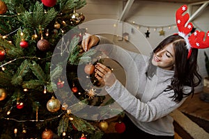 Stylish young woman decorating christmas tree with shiny golden bauble in modern room. Happy girl in reindeer antlers preparing