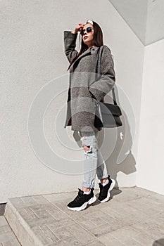 Stylish young woman in fashionable casual youth coat with handbag in sneakers with bandana in sunglasses is resting on sunny day