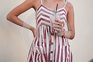 Stylish young woman in a fashionable pink sundress holds a delicious strawberry milkshake in hand. Trendy girl drinks a cool drink